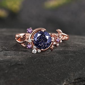 May include: A rose gold ring featuring a round, dark blue, glittery sandstone center stone. The band is shaped like a crescent moon and vine, accented with small, clear, and purple round-cut stones arranged along the sides of the setting.