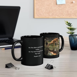 May include: A black ceramic mug featuring a photographic print of a deer drinking from a stream and the white text: As the deer pants for the water, so my soul longs for you My God Psalm 42:1. The mug has a curved handle and a glossy finish.
