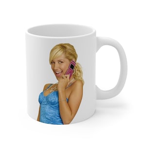 Könnte beinhalten: Weißer Keramikbecher mit einer lächelnden blonden Frau, die ein pinkfarbenes Mobiltelefon hält. Die Frau trägt ein blaues Oberteil und eine Halskette. Der Becher hat einen weißen Griff und ein weißes Inneres.