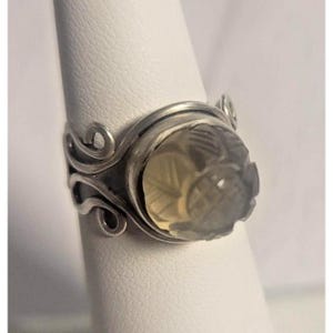 May include: A silver ring with a large, carved, oval-shaped gemstone. The gemstone has a light yellow hue and is intricately carved with a floral design. The ring's band features decorative, swirling silver accents on the sides.