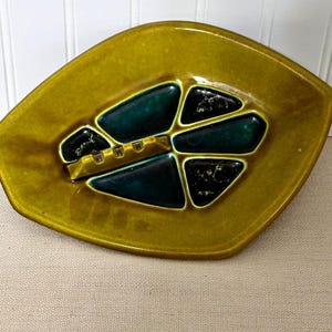 May include: A vintage ashtray with a leaf-like shape. The ashtray is a mustard yellow color with dark teal and black geometric accents. It features a built-in rest for cigarettes.