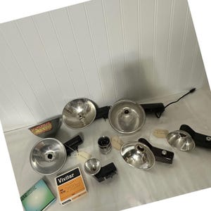 May include: A collection of vintage camera flash units and accessories. The image shows several silver reflector bowls, black flash units, a small metal bowl, a lens, and a box labeled "Vivitar". Some items have attached tags.