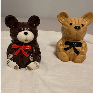 May include: Two ceramic teddy bear figurines. One is brown with a red bow, the other is tan with a black bow. Both have black eyes and noses, and white faces and paws. The bears are sitting upright.