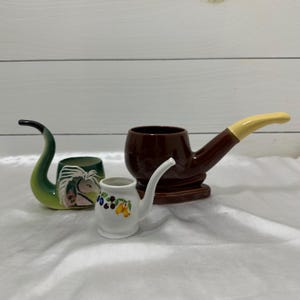 May include: Three ceramic tobacco pipes in different styles. One is green with a curved stem and a horse head design. Another is brown with a yellow mouthpiece. The third is white with a floral design.