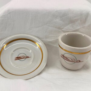 May include: A white ceramic cup and matching saucer set featuring a gold rim and a central graphic of a stylized brown bird in flight within a thin green circle. The design is consistent on both the cup and the saucer, showcasing a clean finish.