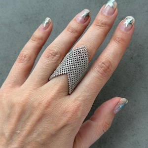 May include: A silver-toned ring featuring a wide, V-shaped design with a fine, textured mesh pattern. The ring covers a significant portion of the finger, showcasing a pointed top edge and a metallic finish that reflects light across its surface.