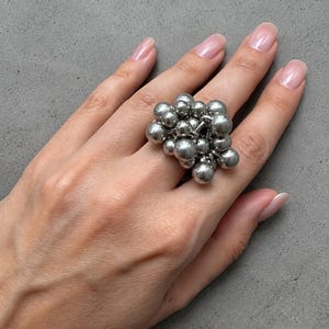 May include: A silver-toned ring featuring a cluster of multiple smooth, spherical metal beads of varying sizes. The beads are arranged in a dense, rounded formation that sits atop the finger, creating a textured, metallic, and dimensional appearance.