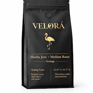 Könnte beinhalten: Schwarzer Kaffeepackung mit goldenem Text und einem Flamingo-Logo. Auf der Packung steht "VELORÁ", "Mocha Java - Medium Roast" und "Heritage". Verkostungsnotizen umfassen gerösteten Kakao, dunkle Kirsche und weiche Erde.