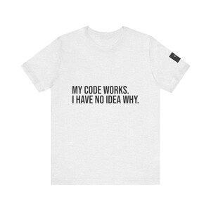 Peut inclure: Un t-shirt gris clair à manches courtes avec le texte noir "MY CODE WORKS. I HAVE NO IDEA WHY." imprimé sur le devant. Une petite étiquette rectangulaire noire est visible sur le bord de la manche gauche, affichant une lettre blanche partielle.