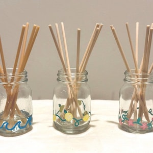 May include: Three clear glass jars filled with wooden diffuser sticks. Each jar is decorated with a different design: blue waves, yellow lemons, and pink flowers. The jars are arranged on a white surface.