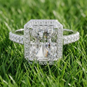 May include: A radiant-cut diamond engagement ring set on a bed of green grass. The ring features a large, rectangular diamond surrounded by a halo of smaller stones. The band is also encrusted with diamonds, creating a sparkling effect.