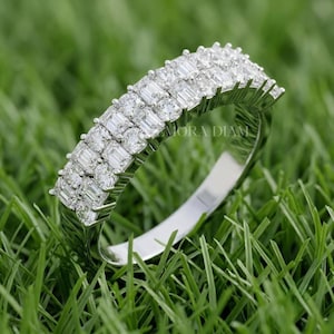 May include: A silver-toned ring featuring two parallel rows of rectangular baguette-cut stones set in a metallic band. The jewelry piece displays a clear, faceted design with a polished finish, showcasing a repeating pattern of stones along the top.