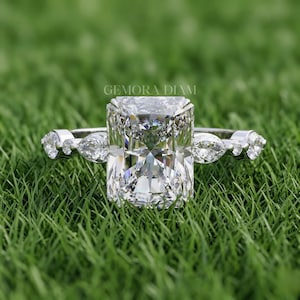 Può includere: Un anello di fidanzamento in argento con un grande diamante taglio radiant e pietre laterali a forma di pera. L'anello è appoggiato su erba verde. Il testo "GEMORA DIAM" è visibile.