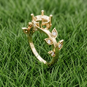 May include: A gold ring setting featuring a central prong mount for a gemstone, accented by leaf-shaped motifs along the band. Each leaf contains a small, round, clear stone. The text GEMORA DIAM is visible on the side of the ring band.