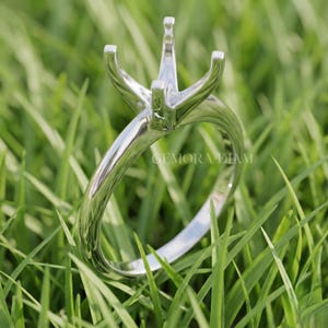 May include: A polished silver-toned metal ring setting featuring a four-prong head designed to hold a gemstone. The band is smooth and rounded, and the metal surface reflects light. The text GEMORA DIAM is visible on the side of the ring.