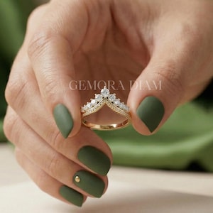 May include: A gold-colored ring with a V-shaped design, adorned with multiple small, clear gemstones. The ring is held against a background of a hand with olive-green nail polish. The text "GEMORA DIAM" is visible.