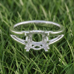 May include: A polished silver-toned metal ring setting featuring a split shank design and a central four-prong mount for a gemstone. The metal surface is reflective and smooth, with the text GEMORA DIAM engraved on the inner band of the ring.