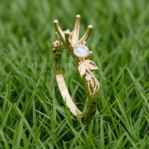 May include: A gold ring featuring a central prong setting for a stone, accented by two small round diamonds and polished gold leaf motifs along the band. The text GEMORA DIAM is visible on the inner surface of the gold band of this jewelry piece.