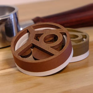 以下が含まれることがあります： 木製のハンドルとステンレス製のカップが付いたコーヒーポルタフィルター。2つの茶色と緑色のコーヒータンパーが前景にあります。タンパーは白いベースと木製のデザインです。