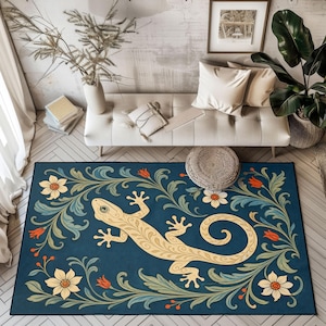 Könnte beinhalten: Ein rechteckiger Teppich mit tiefblauem Hintergrund und einer zentralen, cremefarbenen Eidechsenillustration, die mit komplexen Schnörkelmustern verziert ist. Der Rand ist mit stilisierten cremefarbenen Blumen, grünem Laub und kleinen orangefarbenen Knospen geschmückt.