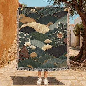 May include: A rectangular woven throw blanket featuring a dark green, gold, and cream landscape pattern with stylized fan-shaped trees, circular floral motifs, and fringed edges, held up to display the intricate, multi-colored textile design.