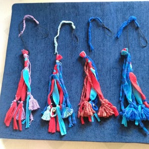 Puede incluir: Cuatro borlas de tela hechas a mano en tonos de rojo, azul y morado, con lazos negros en la parte superior. Las borlas están dispuestas sobre un fondo de tela azul oscuro. Cada borla tiene un diseño único.