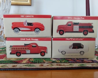 May include: Four vintage car models in boxes. The boxes are red and white with the car's year and model name printed on them. The models include a red 1957 Corvette, a red 1954 Ahrens-Fox fire truck, a red 1948 Task Master truck, and a white 1964 Mustang.