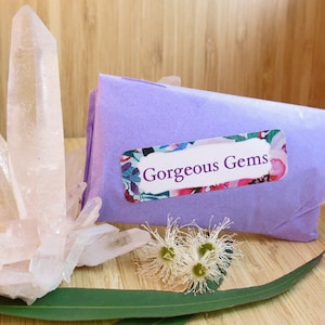 May include: A cluster of clear quartz crystals on a wooden surface with a purple gift bag with a floral label that says "Gorgeous Gems".