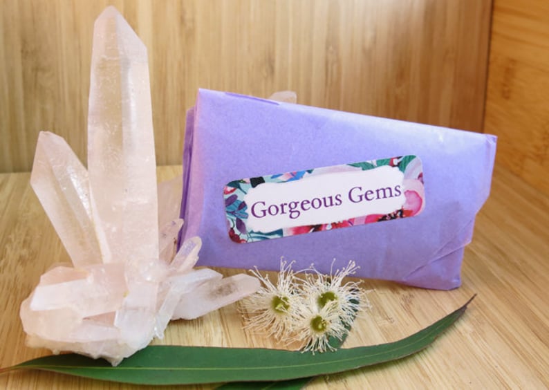 May include: A cluster of clear quartz crystals on a wooden surface with a purple gift bag with a floral label that says "Gorgeous Gems".