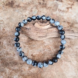 May include: A bracelet made of round snowflake obsidian beads. The beads alternate between black and grey with white speckles. The bracelet is displayed on a natural wooden surface. The beads are approximately 0.8 cm in diameter.