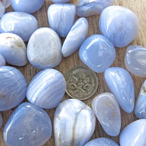 May include: A collection of blue agate tumbled stones, some with white banding, arranged on a wooden surface. A single Australian coin is included for size reference.