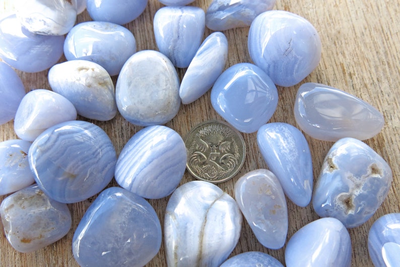 May include: A collection of blue agate tumbled stones, some with white banding, arranged on a wooden surface. A single Australian coin is included for size reference.