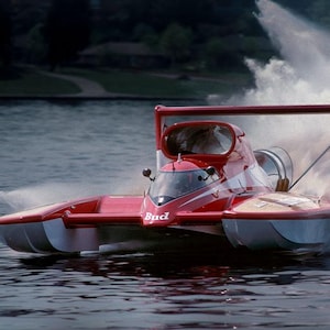Op de afbeelding: Een rode en witte hydroplane boot scheurt over het water en creëert een grote splash. De boot heeft een gestroomlijnd ontwerp met een cockpit en het woord "Bud" aan de zijkant. De achtergrond toont een donkere kustlijn en een bewolkte lucht.