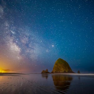 May include: A nighttime landscape featuring a beach, ocean, and a prominent rock formation under a starry sky. The Milky Way is visible, with a gradient of colors from orange to deep blue. The water reflects the sky.