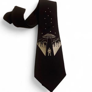 Peut inclure: Cravate noire avec un motif doré d'un OVNI projetant un faisceau sur une silhouette dans une forêt. La cravate a une extrémité pointue et un haut bouclé. Le motif comprend des étoiles dans le ciel nocturne.