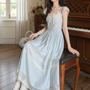 Puede incluir: Un vestido azul claro con ribete de encaje blanco y escote cuadrado. El vestido tiene mangas cortas y una falda fluida. Una cinta blanca está atada en la cintura. El vestido se usa con calcetines blancos. Un piano y flores están en el fondo.