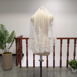 May include: A sheer white bridal veil with delicate floral embroidery along the lower edge. The veil is displayed on a mannequin, showcasing its flowing design. The veil is a bridal accessory.