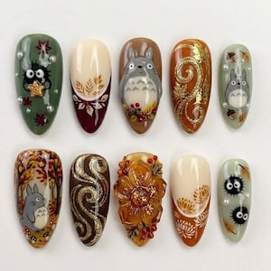 May include: A set of ten almond-shaped press-on nails featuring autumn-themed designs. The collection includes illustrations of grey Totoro characters, black soot sprites, gold glitter swirls, 3D amber flowers, white pearls, and fall leaves.