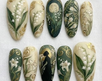 May include: A set of ten almond-shaped nail art designs. Each nail features a different design, including lily of the valley flowers, gold accents, and a silhouette of a woman. The color palette includes cream, green, gold, and black.