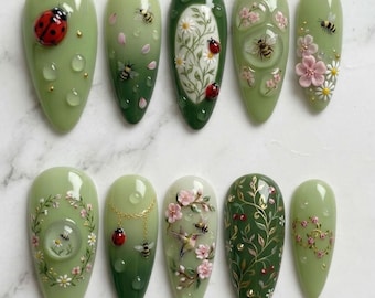 May include: A set of ten green and white artificial nails with various designs. The designs include ladybugs, bees, flowers, and water droplets. Some nails feature gold accents and detailed floral patterns, creating a spring or summer theme.