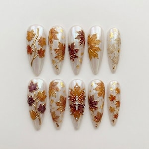 May include: A set of ten almond-shaped press-on nails with a pearlescent white base. The designs feature raised gold-outlined maple leaves in shades of orange, red, and brown, accented with gold foil flakes and small clear rhinestones on one nail.