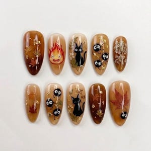 May include: A set of ten almond-shaped press-on nails featuring a warm brown gradient base. Designs include hand-painted black cats, soot sprites, a fire spirit, pressed autumn leaves, and delicate white floral accents with gold foil flakes.