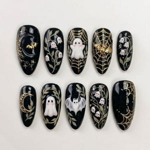 May include: A set of ten almond-shaped press-on nails with a glossy black base. The designs feature raised 3D elements including white ghosts, gold crescent moons, gold spider webs, gold bats, and delicate white lily of the valley floral motifs.