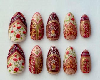 May include: Set of ten almond-shaped artificial nails. Each nail is painted with intricate designs in red, gold, and cream. Floral and ornamental patterns are featured, with a glossy finish. The nails are arranged on a white surface.