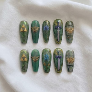 May include: A set of ten green and gold press-on nails with various designs. The designs include the Triforce symbol, a Hylian crest, a blue flower, a sword, and a stone-like pattern. The nails are arranged on a white surface.