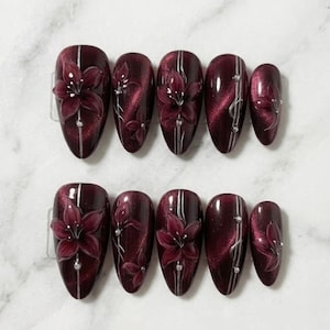 May include: Set of ten burgundy-coloured artificial nails. Each nail features a 3D floral design with silver accents and a vertical silver stripe. The nails are oval-shaped and have a glossy finish.