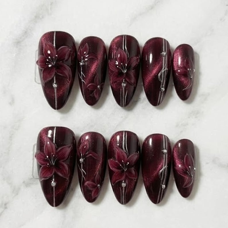 May include: Set of ten burgundy-coloured artificial nails. Each nail features a 3D floral design with silver accents and a vertical silver stripe. The nails are oval-shaped and have a glossy finish.