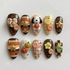 May include: A set of ten almond-shaped press-on nails featuring a brown, orange, and cream colour palette. The designs include Snoopy and Woodstock characters, 3D pumpkin charms, star accents, plaid patterns, and small plastic buttons.