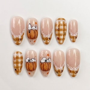 May include: A set of ten almond-shaped press-on nails featuring a nude base with a subtle shimmer. Designs include a brown and white gingham pattern, white lace-style accents, and illustrations of Snoopy resting on an orange pumpkin with autumn leaves.