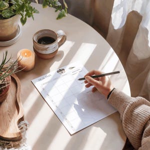 May include: A meal plan notepad with a grid layout, a coffee mug, a lit candle, and a potted plant on a light-colored table. A hand is holding a pen, ready to write on the notepad. The notepad has the words "MEAL PLAN" printed on it.